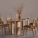 A natural caramel solid wood dining table with an oval top and sculptural pillar legs - Modera Furnish.