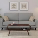 A 35-inch teak-finished solid Sheesham wood coffee table featuring curved waterfall edges, splayed tapered legs, and dual open storage compartments - Modera Furnish.